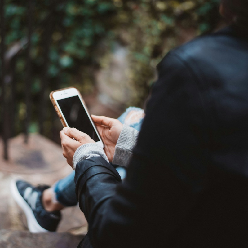 A person holding a smartphone outdoors, wearing a black jacket and ripped jeans.