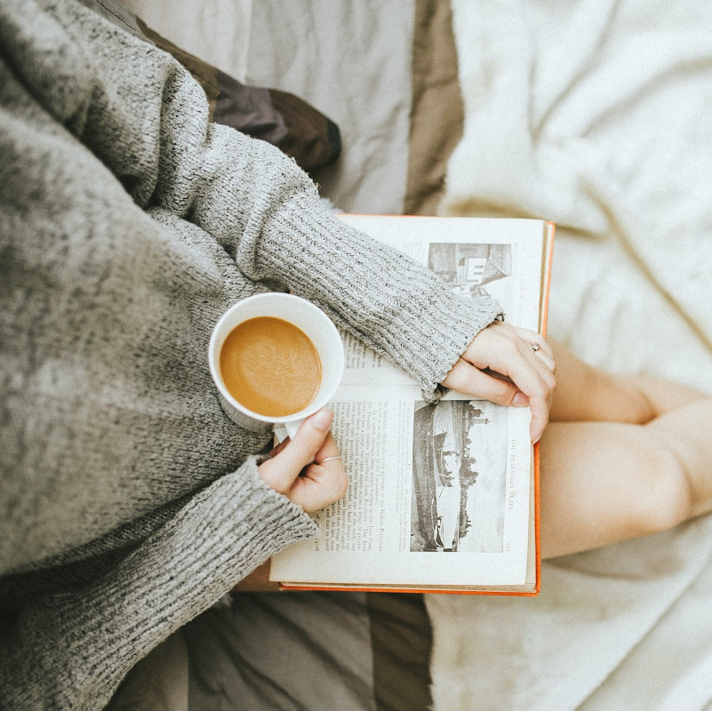 A person in a cozy sweater holding a cup of coffee and reading a book while sitting on a bed.